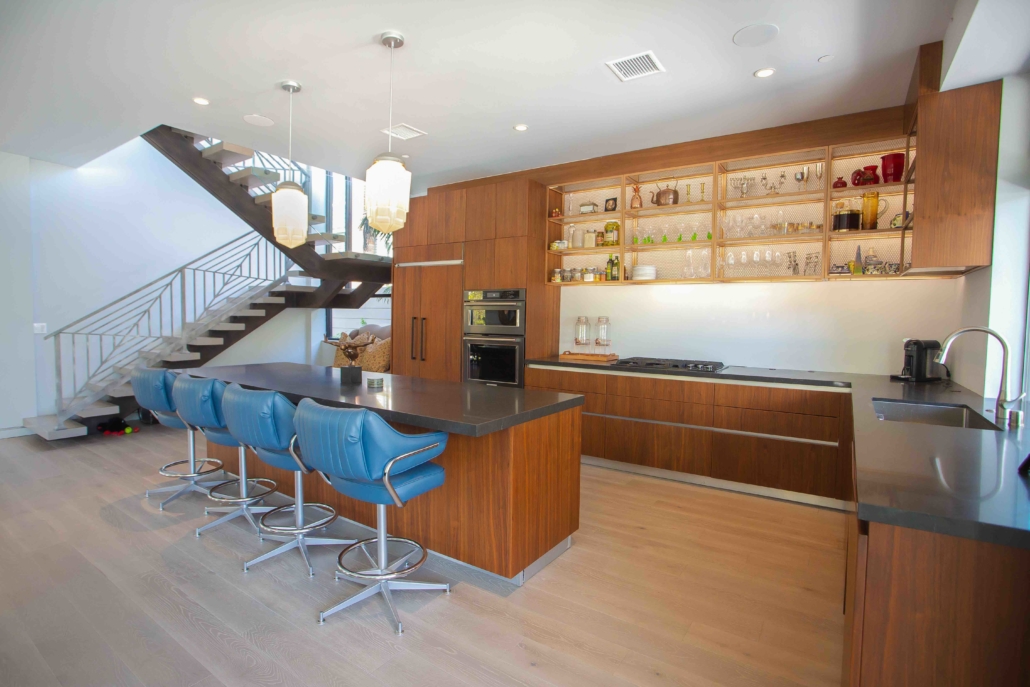 Home bar with wood cabinetry, illuminated shelves, a dark countertop and blue stools